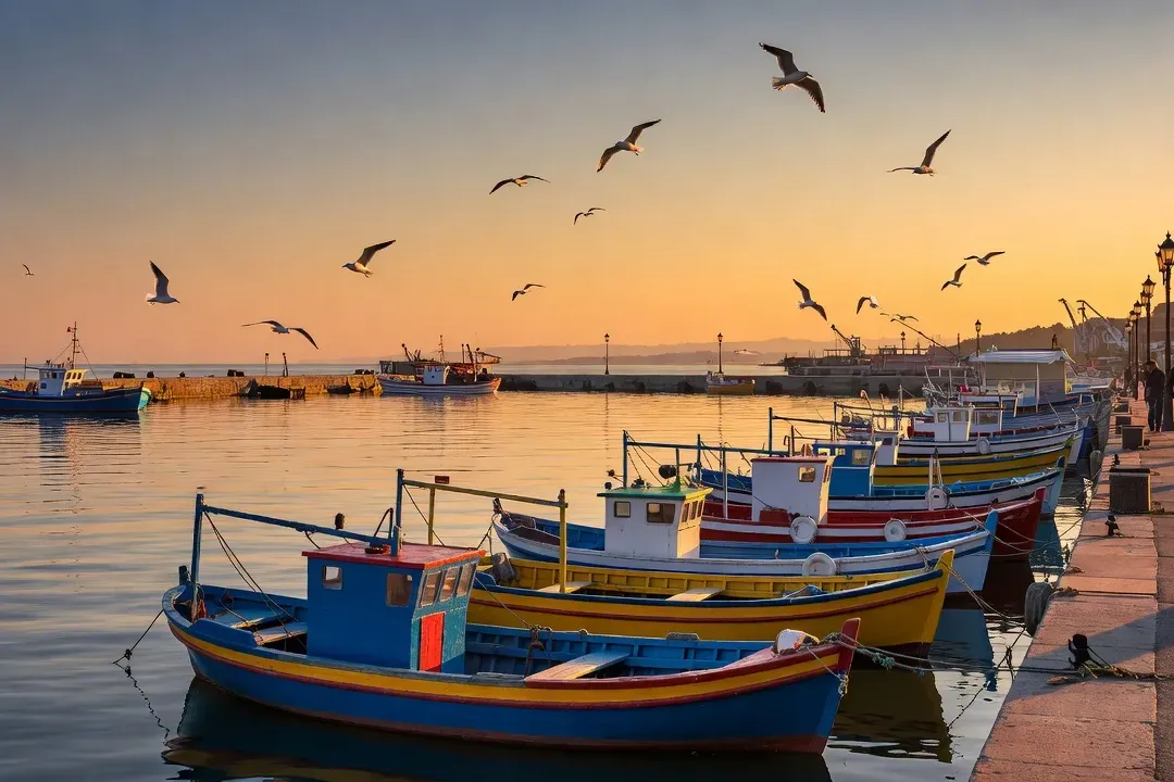 Port de pêche de Bourgas au coucher du soleil, bateaux colorés amarrés et reflets orangés sur l'eau calme